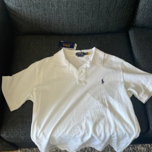 Vit piké t-shirt från Polo Ralph Lauren - Säljer en klassisk vit piké t-shirt från Polo Ralph Lauren med blå broderad logga på bröstet. Modellen är custom slim fit och har korta ärmar samt krage med knappar. Perfekt för en stilren och tidlös look. Pikén är i nytt skick och är bara testad en gång!!