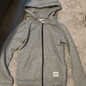 Grå hoodie från Andy by Frank Dandy - Grå hoodie med dragkedja från Andy by Frank Dandy. Tröjan har huva, två fickor framtill och ribbade muddar vid ärmslut och nederkant. Perfekt för en avslappnad stil.