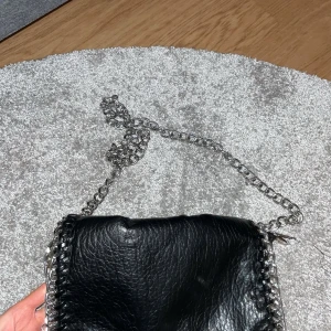 Svart axelväska med kedjedetaljer från Tiamo - Säljer en svart axelväska från Tiamo i skinnimitation med grova silverfärgade kedjor längs kanterna och som axelrem. Väskan har dragkedja upptill och ett stilrent, modernt utseende. Perfekt för att lyfta vilken outfit som helst.