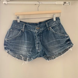 Blå jeansshorts med fransar från PSN och Låg midja - Säljer ett par blå jeansshorts från PSN med råa fransade kanter och klassisk femficksdesign. Shortsen har knappgylf och bakfickor med lock och knapp. Perfekta för varma dagar.