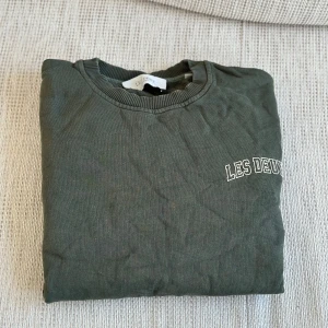 Grön sweatshirt från Les Deux - Mörkgrön sweatshirt från Les Deux med rund halsringning och diskret vit logga på bröstet. Tröjan har ribbade muddar vid halsen och är perfekt för en avslappnad stil.