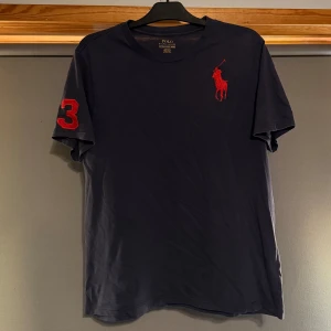 Mörkblå t-shirt från Polo Ralph Lauren - Säljer en mörkblå t-shirt från Polo Ralph Lauren med röd broderad logga på bröstet och röd siffra 3 på ärmen. Klassisk passform och kortärmad modell, perfekt för en stilren look.