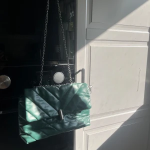 Grön quiltad axelväska med kedja - Säljer en snygg grön quiltad axelväska med silverfärgad kedjerem och metallspänne. Väskan har en modern design och passar perfekt för att lyfta din outfit. Rymmer det viktigaste och är enkel att bära över axeln.