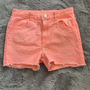 Korallrosa jeansshorts från H&M - Säljer ett par korallrosa jeansshorts från H&M med fransig kant nertill. Shortsen har klassisk femficksdesign och knappgylf. Perfekta för varma dagar och ger en färgklick till outfiten.