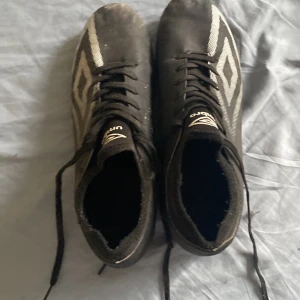 Svarta Umbro fotbollsskor med snörning - Säljer ett par svarta fotbollsskor från Umbro med vita detaljer och snörning. Skorna har klassisk rund tå och dobbar för bra grepp på planen. Perfekta för dig som vill ha stilrena och funktionella skor till match eller träning.