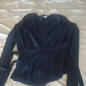 Toppar - Snygg mörkblå omlottopp från Nelly med långa ärmar och knytband i midjan. Toppen har en djup v-ringning och är gjord i ett lätt glansigt, ribbat material som ger en cool struktur. Perfekt för dig som gillar stilrena och trendiga plagg.