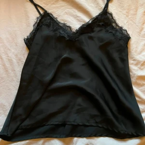 Svart linnetopp med spetsdetaljer - Snygg svart linnetopp med tunna axelband och v-ringning. Toppen har fina spetsdetaljer längs urringningen som ger en extra touch. Perfekt att styla lager på lager eller bära som den är. Mjuk och glansig känsla. Storlek M från bikbok använd fåtal gånger !🙌💕
