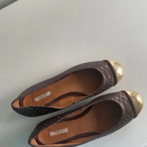 Bruna ballerinaskor med guldtå från Geox - Snygga ballerinaskor från Geox i mörkbrunt skinn med ormskinnsmönster och metallic guld-detalj på tån. Skorna har rund tå och platt sula, perfekt för dig som vill ha något unikt och stilrent. Insidan är i mjukt läder för extra komfort. Säljer pågrund av att de är alldeles för små och har bara legat i garderoben. Storlek 38. Priset går att diskuteras.