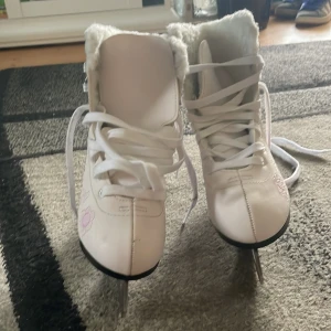 Vita konståkningsskridskor Hudora 35 - Snygga vita konståkningsskridskor från Hudora med ljusrosa broderade blommor på sidorna. Skorna har snörning, fluffigt vitt foder och svart klack. Perfekta för dig som vill sticka ut på isen med stil och komfort.