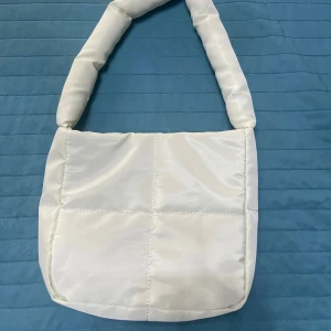 Vit quiltad axelväska  - Snygg vit axelväska  med quiltad design och bred, vadderad axelrem. Väskan har magnetlås i guld.Enkel och stilren modell som rymmer det viktigaste. Mått ungefär 23*19.
