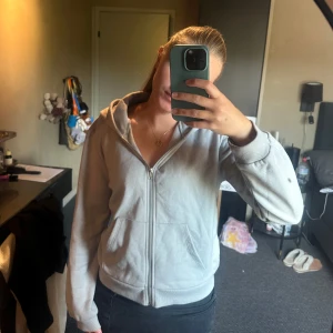 Ljusgrå hoodie med dragkedja - Säljer en ljusgrå hoodie med dragkedja och huva. Tröjan har två fickor framtill och ribbade muddar vid ärmslut och nederkant. Perfekt för chill dagar eller när du vill ha en enkel och stilren look.