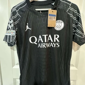 PSG svart matchtröja Marios 11 - Snygg svart PSG fotbollströja med vita detaljer, tryckta loggor och Qatar Airways sponsor på bröstet. Tröjan har korta ärmar, diskreta mönster och vita ränder på axlarna. Namn och nummer Marios 11 på ryggen. Perfekt för dig som älskar fotboll och PSG.