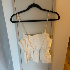 Vit volangig linnetopp med smala band - Supersöt vit linnetopp med smala axelband och volangig, rynkad design. Toppen har en luftig och lätt känsla, perfekt för varma dagar. Snygg att styla med jeans eller kjol för en trendig look.