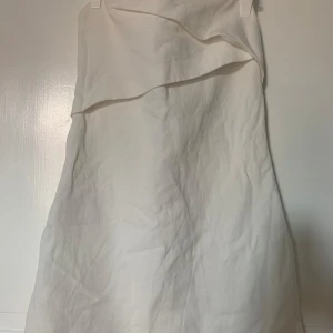 Vit axelbandslös kortklänning - Snygg vit axelbandslös klänning med asymmetrisk volang upptill. Enkel och stilren design som ger en fräsch look. Perfekt för dig som gillar minimalistiskt mode och vill sticka ut med en cool detalj.