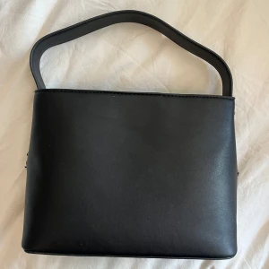 Svart stilren handväska i skinnimitation - Enkel och snygg svart handväska med rak form och bredt handtag. Väskan har en dragkedja på baksidan och silvriga metalldetaljer på sidorna. Perfekt för dig som gillar minimalistisk stil och vill ha en väska som funkar till det mesta. Se defekter på 4:e bilden, fattas knappar på båda sidorna! Har funnits ett långt band till men har tyvärr inte kvar det!❤️