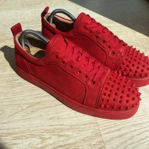 Röda Christian Louboutin sneakers med nitar - Snygga röda sneakers från Christian Louboutin i mocka med matchande röd sula och coola nitar på tån. Skorna har snörning och en platt sula, perfekt för dig som vill sticka ut med en edgy och lyxig stil.