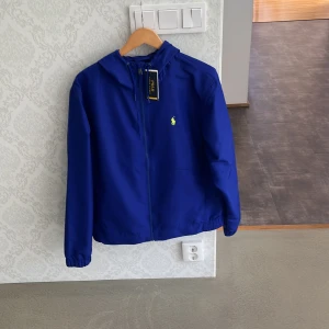 Blå vindjacka från Polo Ralph Lauren - Snygg blå vindjacka från Polo Ralph Lauren med huva och dragkedja framtill. Jackan har elastiska muddar och en gul broderad logga på bröstet. Perfekt för dig som vill ha en stilren och sportig look. Lätt och smidig att bära med sig. Kan gå ner i pris vid snabb affär 
