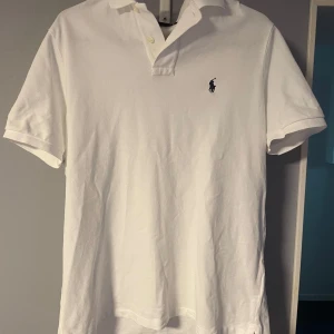 Vit pikétröja från Polo Ralph Lauren - Klassisk vit pikétröja från Polo Ralph Lauren i bomull. Tröjan har korta ärmar, krage med knappar och den ikoniska broderade loggan på bröstet. Perfekt för en clean och stilren look.