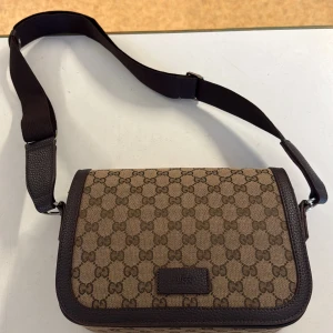 Gucci brun axelväska med GG-mönster - Snygg axelväska från Gucci i beige och brunt med klassiskt GG-mönster. Väskan har justerbar axelrem, detaljer i brunt skinn och dragkedjefack på insidan. Perfekt storlek för det viktigaste och en ikonisk look som lyfter varje outfit.