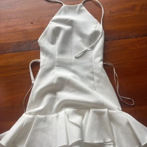 Vit kort klänning med volang och snörning - Supersöt vit kort klänning med tunna axelband och snygg snörning i ryggen. Klänningen har en figurnära passform upptill och en volangkant nertill som ger extra flärd. Perfekt för sommarens alla tillfällen!