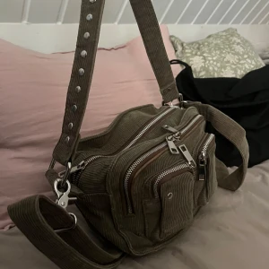 Beige manchesterväska med dragkedjor - Säljer en beige Nunoo axelväska i manchester med bred, justerbar rem och coola silverfärgade nitar. Väskan har flera fack med dragkedjor och en praktisk design som rymmer allt.