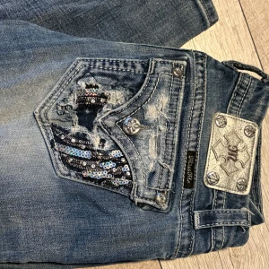 Miss Me jeans med paljettdetaljer - Snygga blå Miss Me jeans med slitningar och paljetter på bakfickan. Jeansen har låg midja, raka ben och coola silverdetaljer. Perfekta för dig som gillar en unik och glittrig look. Materialet är klassisk denim med stretch för extra komfort.