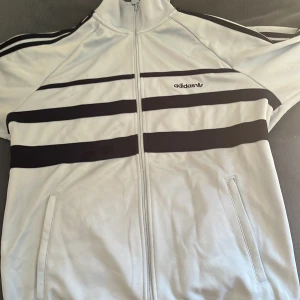 Adidas randig vindjacka ljusblå/mörkblå  - Säljer en snygg Adidas vindjacka i ljusblått med mörkblå ränder  och klassiska stripes på ärmarna. Jackan har dragkedja framtill, ståkrage och två fickor. Perfekt för dig som gillar sportig och retro stil.