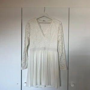 Vit spetsklänning med v-ringning - Superfin vit kortklänning med långärmade spetsärmar och v-ringad hals. Överdelen har broderade blommor och spetsdetaljer, medan kjolen är lätt och flowy. Perfekt för student eller sommarens fester! 