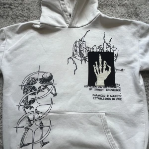 Vit hoodie Paranoid Society med tryck - Vit hoodie från Paranoid Society med stora svarta grafiska tryck fram och bak, bland annat skelett, dödskalle, måne och handmotiv. Hoodien har huva och känguruficka. Materialet är mjuk bomull och passformen är normal. Perfekt för dig som gillar streetwear med edge.