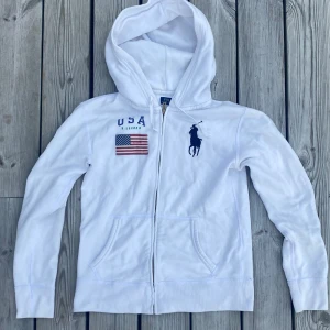 Vit hoodie från Polo Ralph Lauren XS - Vit hoodie från Polo Ralph Lauren med dragkedja, huva och fickor. Framsidan har USA-flagga, blå Polo-logga och texten 'USA'. Skön bomullskvalitet. Rare usa RalphLauren hoodie.