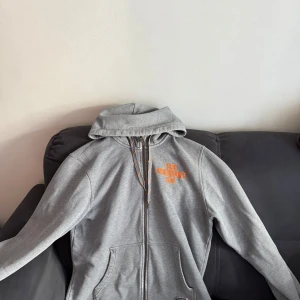Grå hoodie från Peak Performance - Snygg grå hoodie från Peak Performance med orange broderad logga på bröstet. Tröjan har huva med snörning, dragkedja framtill och praktiska fickor. Perfekt för chill dagar eller när du vill ha en sportig look.