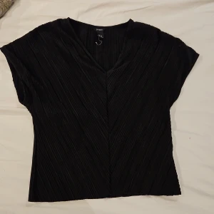 Svart plisserad topp från Lindex - Svart plisserad topp från Lindex i storlek XS. Toppen har v-ringning, korta ärmar och ett snyggt diagonalt veckat tyg som ger en cool struktur. Perfekt för dig som gillar stilrena och trendiga plagg med det lilla extra.