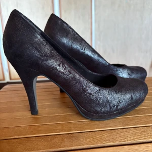 Svarta pumps från Tamaris med klack - Snygga svarta pumps från Tamaris med hög klack och rund tå. Skorna har en diskret glansig yta med struktur och är tillverkade i syntetmaterial. Perfekta för dig som vill ha en klassisk och stilren look.