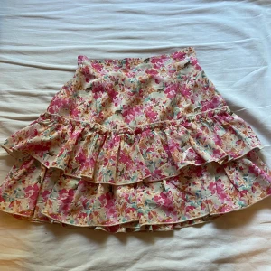 Blommig volangkjol i pastellfärger - Supersöt kort kjol med blommigt mönster i rosa, blått och beige. Kjolen har dubbla volanger som ger extra fladdrig känsla och en romantisk vibe. Perfekt för dig som gillar färg och vill sticka ut lite extra i sommar.