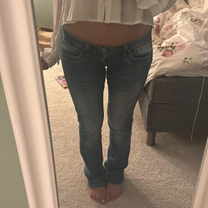 Lågmidjade blå jeans med straight fit - Säljer ett par snygga lågmidjade blå jeans med klassisk femficksdesign och lätt slitna detaljer. Jeansen har en rak passform och är tillverkade i mjukt denimtyg som sitter skönt på kroppen. Perfekta för en chill och avslappnad look.
