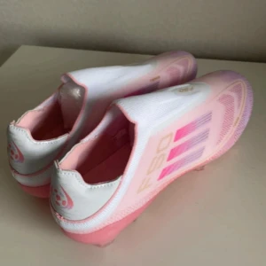 Lamine Yamals Adidas F50 limited edition  - Säljer ett par av lamine yamals Adidas F50  fotbollsskor i en snygg mix av vitt och olika rosa nyanser. Skorna har en strömlinjeformad design utan snörning, med klassiska tre ränder på sidan och F50-logga. Ovandelen är i syntetmaterial och sulan har fasta dobbar för gräsplan.