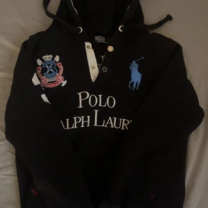 Svart hoodie Polo Ralph Lauren S - Svart hoodie från Polo Ralph Lauren med stora broderade loggor och nummerdetaljer i blått, vitt och rött. Tröjan har huva med snörning, lång ärm och är tillverkad i 100% bomull. Klassisk streetstil med tydliga Polo- och nummerapplikationer fram och bak. Tröjan är ganska unik. Tveka inte på att höra av dig!🤗