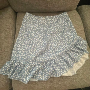 Blå blommig omlottkjol med volang - Supersöt blå kortkjol med vitt blommönster och volangkant. Kjolen har omlottmodell som ger en snygg och feminin look. Perfekt för dig som gillar romantisk och somrig stil!