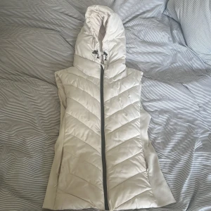 Zara väst - Snygg beige dunväst med huva och dragkedja framtill. Västen har quiltad design med snedställda sömmar och dragsko i huvan. Perfekt för lager-på-lager och ger en clean look till din outfit. Liten sminkfläck men syns knappt🩷