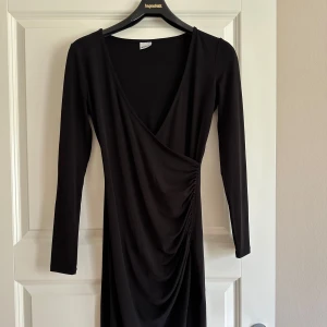 Svart omlott fest nyår klänning drapering Gina Tricot - Snygg svart kortklänning med omlott och draperad detalj framtill. Klänningen har lång ärm och djup v-ringning.