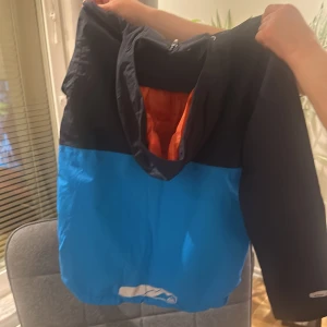 Blå och svart vindjacka (PRISET KAN DISKUTERAS VID SNABB AFFÄR) - Snygg tvåfärgad vindjacka från McKinley i blått och svart med huva och dragkedja framtill. Praktisk bröstficka med dragkedja och elastisk nederkant. Perfekt för blåsiga dagar och utomhusaktiviteter. Ej använd och i nyskick.