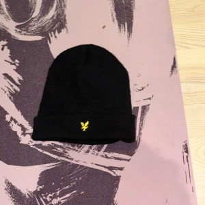 Svart mössa från Lyle & Scott - Svart ribbstickad mössa från Lyle & Scott med klassisk uppvikt kant och gul broderad logga framtill. Enkel och stilren design som passar till det mesta. Perfekt accessoar för kyliga dagar.