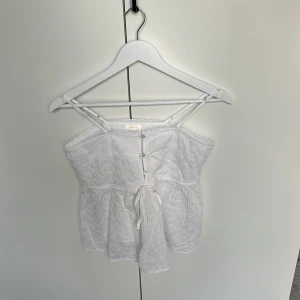 Vit broderad linnetopp Gina Tricot - Supersöt vit linnetopp från Gina Tricot med tunna axelband, broderade blommor och spetskant upptill. Toppen har knappar framtill och en liten knytning i midjan för extra detalj. Perfekt för varma dagar och somriga vibes!