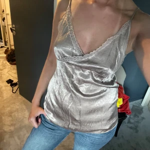 Champagnefärgad linnetopp i satin - Säljer en glansig champagnefärgad linnetopp i satin med smala axelband och omlott-skärning. Toppen har v-ringning med spetsdetaljer längs kanten och en lätt figurnära passform. Perfekt för dig som gillar en stilren men ändå lyxig vibe.