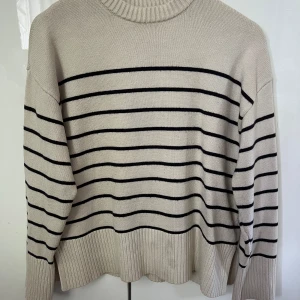 Beige randig stickad tröja från Zara - Snygg beige stickad tröja från Zara i storlek S med svarta horisontella ränder. Tröjan har ribbad mudd vid hals, ärmslut och nederkant. Perfekt för lager-på-lager och chill vibes. Klassisk passform och långärmad modell. Fläck finns på nedre framsida (se sista bild)