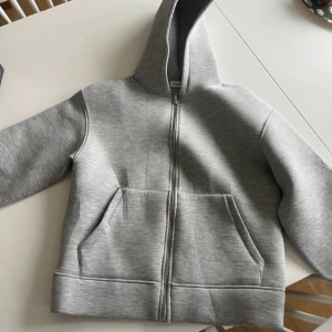 Grå hoodie med dragkedja från Kappahl - En stilren grå hoodie från Kappahl med hel dragkedja framtill och stor huva. Tröjan har två rymliga fickor på magen och är tillverkad i mjuk bomullsblandning. Perfekt för chill dagar eller när du vill ha en enkel och clean look.