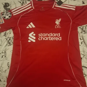 Liverpool matchtröja Wirtz 17 Adidas - Liverpool FCs officiella röda matchtröja från Adidas med tryck Wirtz 17 på ryggen. Tröjan har vita detaljer, klubbmärke och sponsorlogga på bröstet, samt Champions League-märke på ärmen. Tillverkad i lätt funktionsmaterial (AEROREADY) och har klassiska tre vita ränder på axlarna. Tvätta för han så att inte trycket förstörs