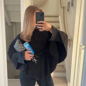Missäy poncho  - Snygg svart kappa med omlottknäppning och bälte i midjan. Jackan har vida ärmar och en minimalistisk design som ger en cool och stilren look. Perfekt för dig som gillar oversized och trendiga siluetter.