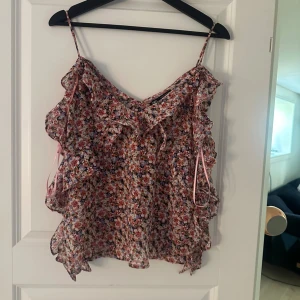 Blommig volangtop från Zara - Supersöt blommig linnetopp från Zara med tunna axelband och volangdetaljer. Toppen har en mix av rosa, lila och vita blommor på en ljus botten och en liten rosett framtill. Perfekt för varma dagar och ger en romantisk vibe.