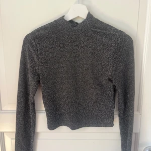 Glittrig silver polotopp med öppen rygg - Säljer en glittrig silverfärgad polotopp med lång ärm och croppad passform. Toppen har en snygg öppen rygg med twistad detalj vid halsen som ger en unik look. Perfekt för dig som vill sticka ut med lite extra glitter.